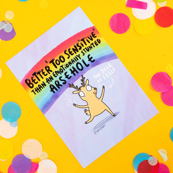 A light lilac postcard with a rainbow background with an illustration of a deer on it with an arrow pointing saying, 'the deer of feels'. Text above reads 'BETTER TOO SENSITIVE THAN AN EMOTIONALLY STUNTED ARSEHOLE' the card is on a yellow background with confetti around it