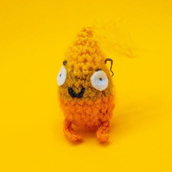 Spark Of Hope Crochet Pal handmade plush character by Katie Abey and Forest Folk Crochet, fluffy orange and yellow crochet creature with wide eyes and tiny legs. Made from vegan friendly yarn and stuffed to keep its shape, inspired by the “Keeping My Tiny Spark Burning” illustration. Quirky handmade crochet gift photographed on a bright yellow background.