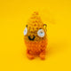 Spark Of Hope Crochet Pal handmade plush character by Katie Abey and Forest Folk Crochet, fluffy orange and yellow crochet creature with wide eyes and tiny legs. Made from vegan friendly yarn and stuffed to keep its shape, inspired by the “Keeping My Tiny Spark Burning” illustration. Quirky handmade crochet gift photographed on a bright yellow background.