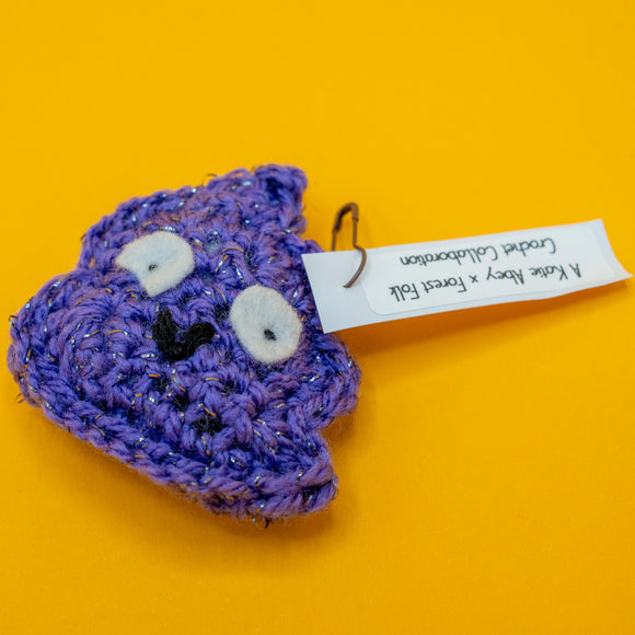 Handmade crochet Galactic Poo character plush by Katie Abey, featuring large felt eyes and a cheeky smile. Made with purple sparkly vegan yarn and lightly stuffed to hold its shape. A small novelty crochet toy created in collaboration with Forest Folk Crochet, perfect as a quirky gift for lovers of weird and whimsical art.