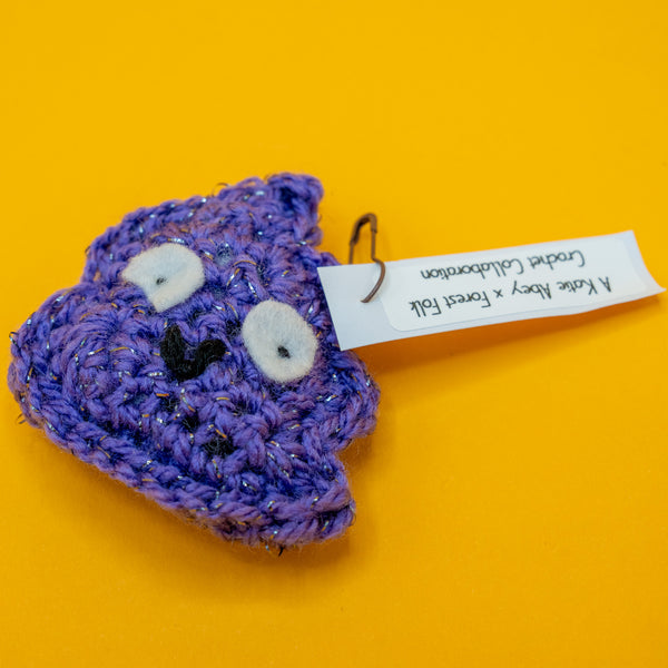 Handmade crochet Galactic Poo character plush by Katie Abey, featuring large felt eyes and a cheeky smile. Made with purple sparkly vegan yarn and lightly stuffed to hold its shape. A small novelty crochet toy created in collaboration with Forest Folk Crochet, perfect as a quirky gift for lovers of weird and whimsical art.