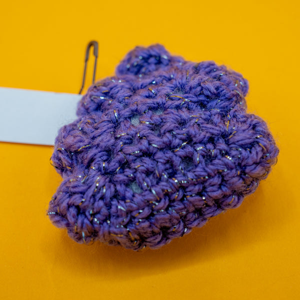 Close up of the handmade Galactic Poo Crochet Pal plush by Katie Abey and Forest Folk Crochet. The small purple crochet toy features sparkly vegan yarn and a soft stuffed shape. A quirky novelty crochet character inspired by Katie Abey’s galactic poo illustration, photographed on a bright yellow background.