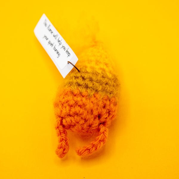 Back view of the Spark Of Hope Crochet Pal handmade character by Katie Abey x Forest Folk Crochet. Small fluffy orange and yellow crochet creature with tiny legs, made using vegan friendly yarn and soft stuffing to hold its shape. Inspired by the “Keeping My Tiny Spark Burning” illustration by Katie Abey, photographed on a bright yellow background as a cute handmade alternative gift.