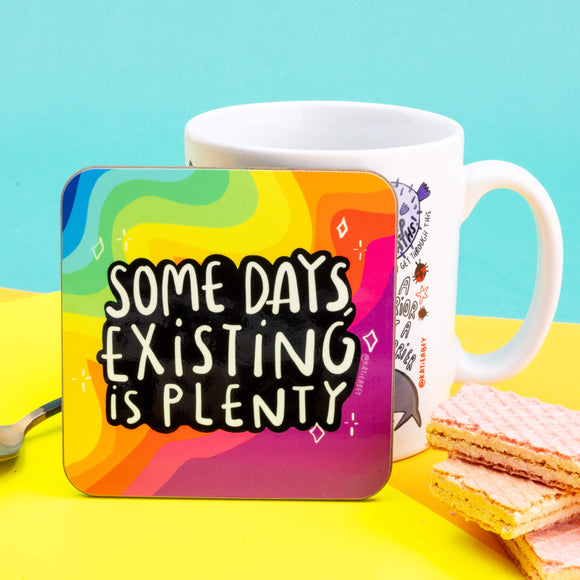 A coaster designed and illustrated by Katie Abey with rainbow background and text saying, 'some days, existing is plenty'