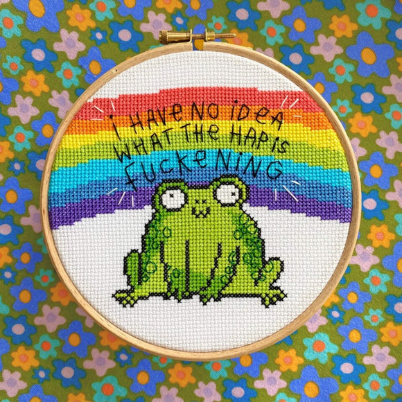 The I Have No Idea Frog Cross Stitch Kits on a green, yellow, blue and pink floral fabric. The white cross stitch fabric with light wood circle frame has a stitched rainbow across the middle with black stitched text reading 'I have no idea what the hap is fuckening' with a cross stitched smiling green frog underneath.