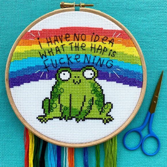 The I Have No Idea Frog Cross Stitch Kits on a blue fabric with the corresponding threads underneath and blue scissors. The white cross stitch fabric with light wood circle frame has a stitched rainbow across the middle with black stitched text reading 'I have no idea what the hap is fuckening' with a cross stitched smiling green frog underneath.