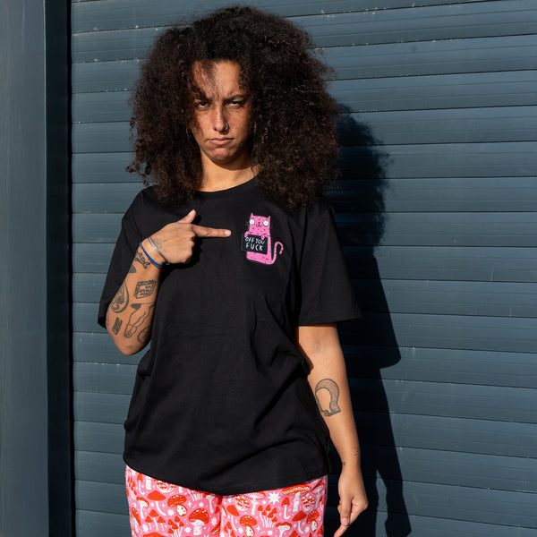 Front view of a model wearing the Sweary Cat Off You F*ck Adults Tee in black by Katie Abey. The model points at the pink illustrated cat graphic on the chest. The black T shirt has a relaxed unisex fit and is styled with pink patterned trousers. Shot outdoors against a blue door in natural light.
