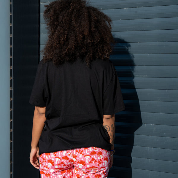 Back view of a model wearing the Sweary Cat Off You F*ck Adults Tee in black by Katie Abey. The relaxed fit black T shirt is paired with pink mushroom print trousers. The illustration is not visible from the back. The model stands outdoors against a blue industrial door, showing the casual drape and length of the tee.
