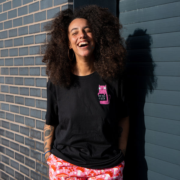 Model laughing while wearing the Sweary Cat Off You F*ck Adults Tee in black by Katie Abey. The pink cartoon cat illustration is clearly visible on the chest of the black T shirt. A relaxed, unisex fit tee photographed outdoors in daylight, styled with colourful trousers for a fun, alternative look.
