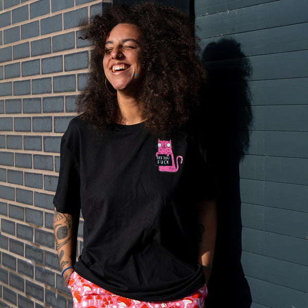 Lifestyle image of a model smiling while wearing the Sweary Cat Off You F*ck Adults Tee in black by Katie Abey. The pink illustrated cat graphic is visible on the chest. The black T shirt has a casual fit and is styled with bright patterned trousers. Photograph taken outdoors against a brick wall and blue door.
