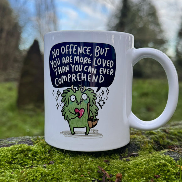 White ceramic mug photographed outdoors on a mossy stone surface, featuring a cute green illustrated character holding a heart and bag with the quote “No offence, but you are more loved than you can ever comprehend”. Designed by Katie Abey, a UK illustrator known for encouraging people to embrace their weirdness. Natural blurred garden background.
