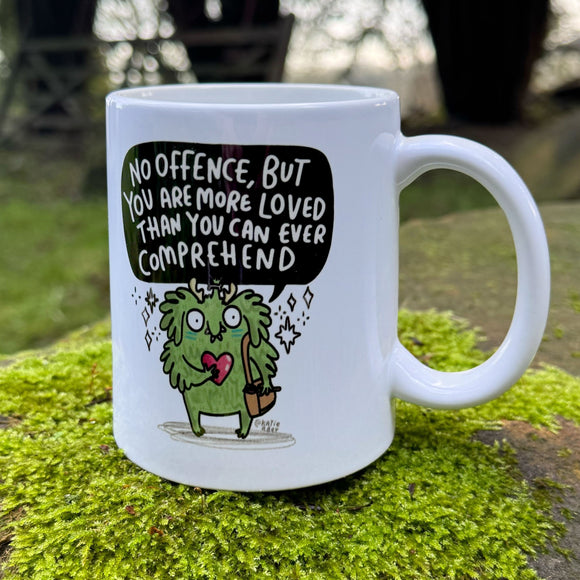 Side view of a white 10oz ceramic mug with handle visible, placed on green moss outdoors. The mug features a hand drawn green character and a positive affirmation reading “No offence, but you are more loved than you can ever comprehend”. Illustrated and printed in the UK by Katie Abey.
