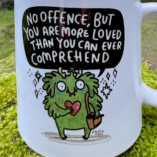 Close up detail of a white ceramic mug showing a whimsical green character illustration holding a heart beneath the quote “No offence, but you are more loved than you can ever comprehend”. Designed by UK illustrator Katie Abey, known for quirky affirmations and encouraging self acceptance.
