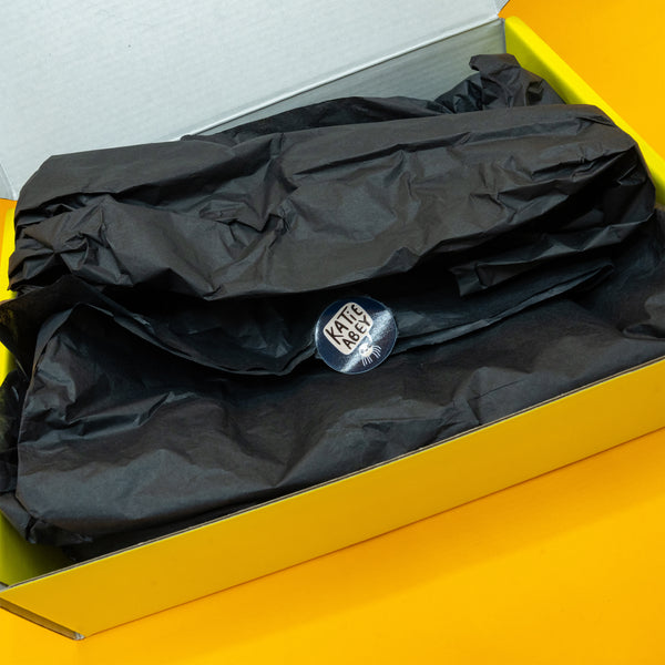 Close up of the inside of the Box of Brilliance by Katie Abey. A yellow box filled with black tissue paper and a katie abey spider circle sticker.
