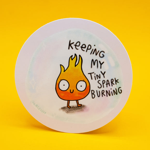 Keeping My Tiny Spark Burning vinyl sticker by Katie Abey featuring a cute smiling flame character with wide eyes and hand drawn lettering. Circular waterproof vinyl sticker on white background. Motivational quirky illustration encouraging self care, resilience and embracing your weirdness. Approx 8cm decorative laptop, journal or water bottle sticker.
