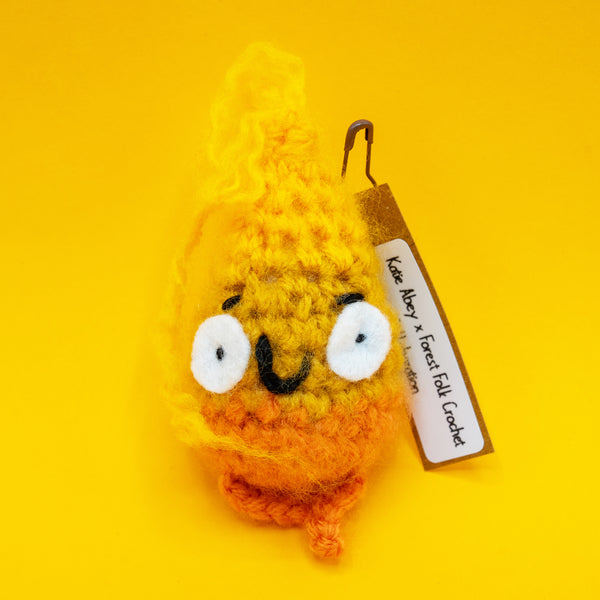 Handmade Spark Of Hope Crochet Pal character by Katie Abey x Forest Folk Crochet, fluffy orange and yellow crochet plush with wide eyes and little legs, made from vegan friendly yarn and stuffed to hold its shape. Cute quirky handmade crochet mascot inspired by Katie Abey’s “Keeping My Tiny Spark Burning” illustration, photographed on a bright yellow background.