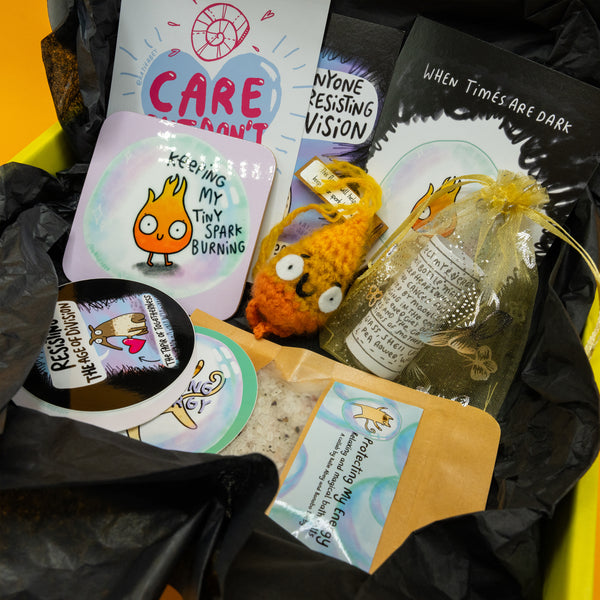 Inside details of the Box of Brilliance by Katie Abey of January's theme, Protect Your Energy. A yellow box full of black tissue paper with the box contents, three postcards with katie abey artwork, a flame artwork coaster, a crochet flame, protect my energy spell bottle, relaxing and magical bath salts and three stickers. 