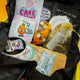 Inside details of the Box of Brilliance by Katie Abey of January's theme, Protect Your Energy. A yellow box full of black tissue paper with the box contents, three postcards with katie abey artwork, a flame artwork coaster, a crochet flame, protect my energy spell bottle, relaxing and magical bath salts and three stickers. 