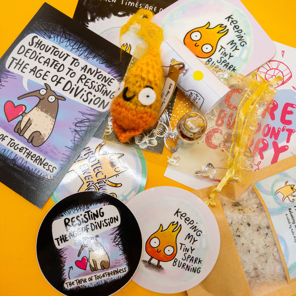 Contents of the Box of Brilliance by Katie Abey of January's theme, Protect Your Energy, three postcards with katie abey artwork, a flame artwork coaster, a crochet flame, protect my energy spell bottle, relaxing and magical bath salts and three stickers. 