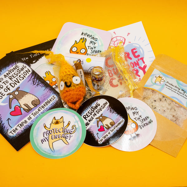 Contents of the Box of Brilliance by Katie Abey of January's theme, Protect Your Energy, three postcards with katie abey artwork, a flame artwork coaster, a crochet flame, protect my energy spell bottle, relaxing and magical bath salts and three stickers. 