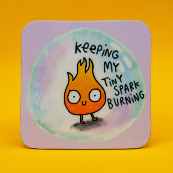 A pastel purple coaster with an illustrated design of an iridescent bubble, a tiny smiling orange flame, and white sparkles, with black text reading 'keeping my tiny spark burning'. An illustrated design by Katie Abey. Square coaster with rounded corners, motivational illustrated design, made in Britain and shown on a yellow background. Quirky positive gift for friends, mental health encouragement, colourful desk or coffee table accessory.