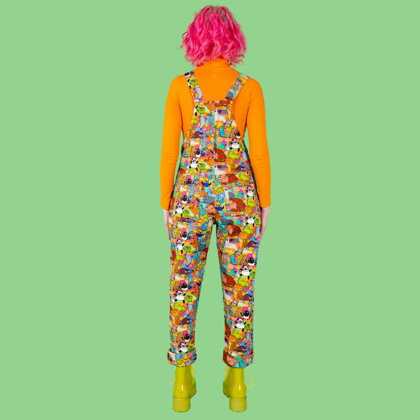 Lottie a pink hair tall female white model is standing with her arms out wearing a pair of dungarees with characters by Katie Abey all over them. She is smiling and looking away from camera. She has an orange roll neck and green boots on and is against a light green background.