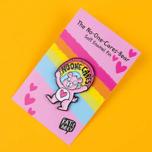 This Katie Abey "No-One Cares Bear" pin features a glittery pink bear with a heart on its belly, holding a rainbow banner that says "No-One Cares." The pin is tilted slightly, giving it a playful, sassy edge, and is displayed on a pink backing card with rainbow stripes and hearts. Ideal for adding some quirky humour to your jacket, bag, or pin collection, this soft enamel pin captures Katie Abey’s distinctive weird, bold style. A fun, unique gift for those who love to stand out.