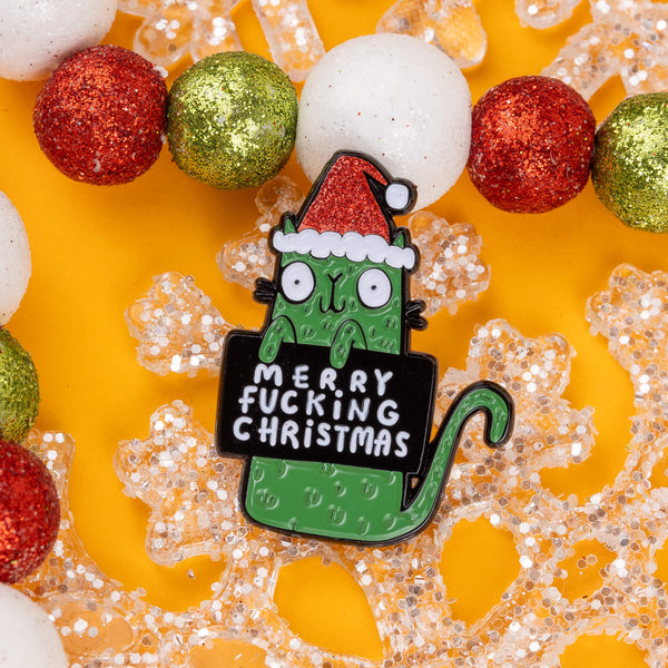 A quirky enamel pin by Katie Abey featuring a green cat in a Santa hat, holding a sign that says "Merry F***ing Christmas." The pin is surrounded by festive, glittery red, green, and white baubles, perfect for adding a touch of weirdness to your holiday attire. Katie Abey is known for unique and sassy designs, encouraging you to embrace your inner oddball this festive season. Ideal for gifting or personal style. Bright yellow background adds an extra pop of colour.