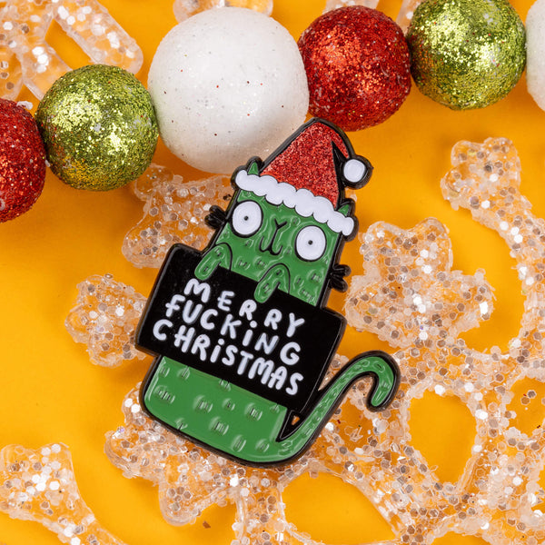 A fun and sassy Christmas enamel pin by Katie Abey featuring a green cat with bulging eyes and a Santa hat. The pin reads "Merry F***ing Christmas," and is surrounded by red, green, and white glittery baubles on a bright yellow background. Known for quirky, humorous designs, Katie Abey inspires you to celebrate your weird side this festive season. This pin is an ideal gift or a quirky addition to your holiday outfit.