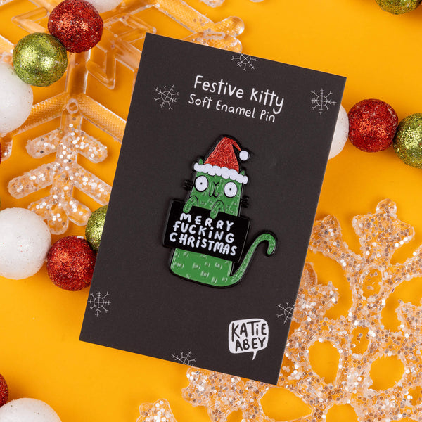 Katie Abey's festive enamel pin features a green cat in a Santa hat, holding a cheeky sign that says "Merry F***ing Christmas." The pin is displayed on a black backing card, framed by glittery baubles and snowflakes on a bright yellow background. Katie Abey, known for her quirky designs, encourages you to embrace your weirdness this Christmas. Perfect as a fun gift or to add some cheeky holiday spirit to your outfit, this pin is a must-have for anyone with a sassy side.