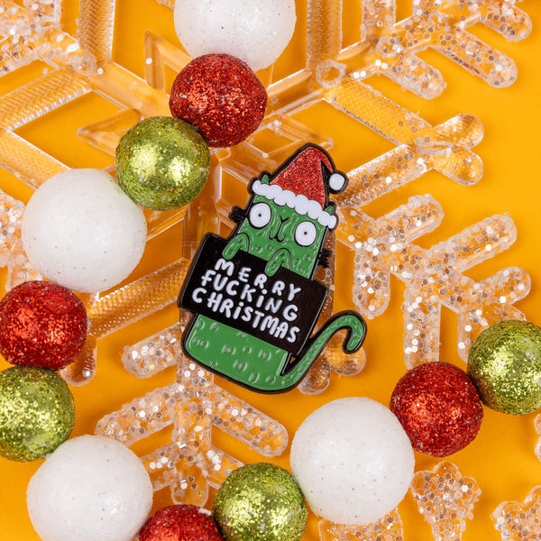 This bold and cheeky enamel pin by Katie Abey features a green, wide-eyed cat wearing a Santa hat and holding a sign that reads "Merry F***ing Christmas." Surrounded by glittery red, white, and green baubles, this pin adds a touch of quirky holiday charm. Katie Abey's designs encourage you to embrace your inner weirdo. Perfect for anyone with a sarcastic streak this Christmas. The pin pops against a bright yellow backdrop, making it an eye-catching accessory or gift.