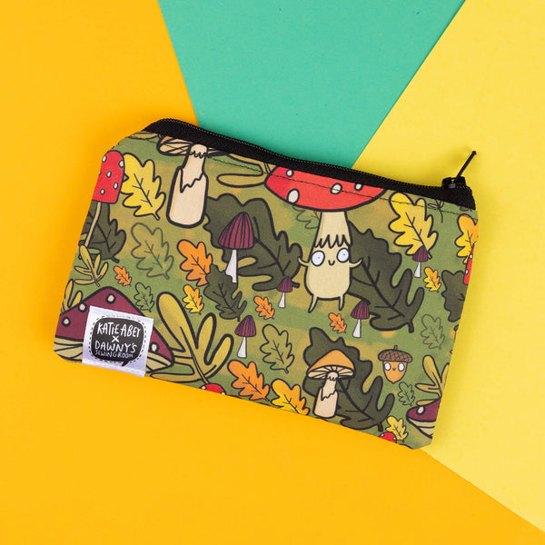 This Katie Abey coin purse features her illustrated mushroom characters, set against a vibrant forest-inspired backdrop of toadstools, acorns, and autumn leaves. Perfect for keeping your coins or small items safe while showing off your love for quirky accessories. The green-toned design is ideal for fans of weird and wonderful patterns, and the purse is finished with a collaborative tag from Katie Abey x Dawny's Sewing Room, showcasing its handmade charm.