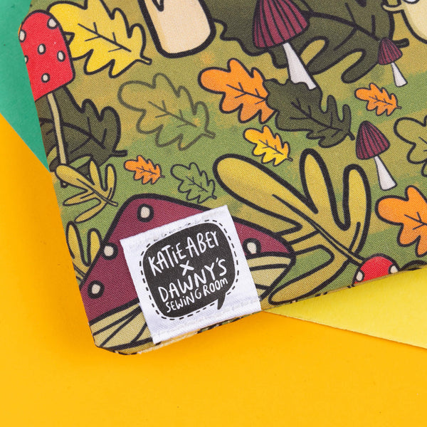 Close up of the mushroom bois coin purse features her illustrated mushroom characters, set against a vibrant forest-inspired backdrop of toadstools, acorns, and autumn leaves. Perfect for keeping your coins or small items safe while showing off your love for quirky accessories. The green-toned design is ideal for fans of weird and wonderful patterns, and the purse is finished with a collaborative tag from Katie Abey x Dawny's Sewing Room, showcasing its handmade charm.