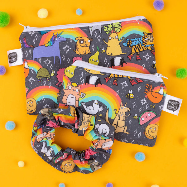 Katie Abey pouch set and matching scrunchie with a quirky rainbow animal print. The pouch features adorable illustrations like a fox, unicorn, and sloth with pops of vibrant colours against a black background. Made with eco-friendly materials, the set has white zip closures for secure storage, with two pouch sizes for different uses. The matching scrunchie ties the look together, perfect for bold, playful style. Photographed on a yellow background with colourful pompoms scattered.