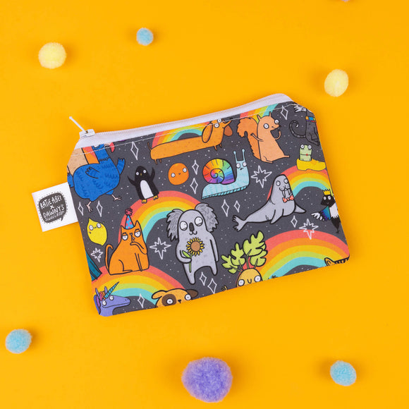 Katie Abey’s rainbow animal coin pouch with a colourful print featuring a sloth, orange cat, blue unicorn, and koala, all embracing their weirdness under a rainbow. Set against a black background with fun details like stars and white zip closure. Perfect for carrying small essentials with style. Photographed on a yellow background with scattered pom-poms for added quirkiness.