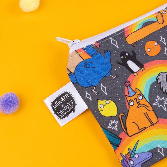 Close-up of Katie Abey pouch with a rainbow animal print, featuring an orange cat in a party hat, a blue bird, and a penguin surrounded by stars and rainbows. The quirky characters are printed on a black background with a white zip closure. Photographed against a yellow background with colourful pom-poms. Ideal for those who love fun, weird accessories.