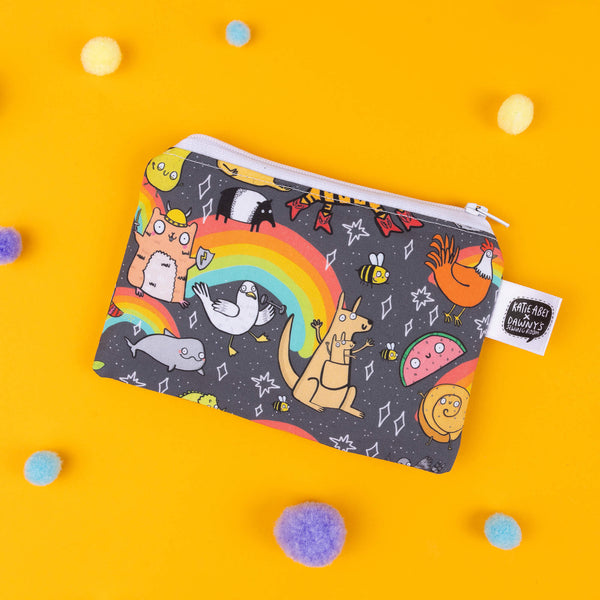 Katie Abey's small pouch featuring a playful rainbow animal print. This quirky accessory showcases an array of fun characters like a fox, kangaroo, and pigeon under a vibrant rainbow on a black background with stars. Complete with a white zip closure, this pouch is perfect for keeping essentials in one place while embracing your weird side. Photographed on a bright yellow background with scattered pom-poms for added whimsy.