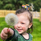 A child with blonde hair styled in a small ponytail, held in place with a Katie Abey rainbow positivity scrunchie, featuring colourful quirky characters like a unicorn and fox. The child smiles joyfully while holding a dandelion, standing in a green outdoor setting. The scrunchie adds a fun pop of colour, and the whimsical design matches the playful, carefree moment. The background has a magical, dreamy atmosphere, perfect for celebrating Katie Abey's embrace of weirdness and fun.