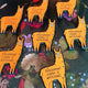 The Judgement Llama Magnet laying on a foliage desk amongst multiple copies of the same magnet. The yellow llama shaped magnet has a scowled expression with black text across its body reading 'judgement llama is watching'.