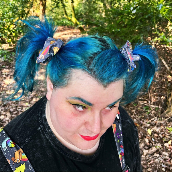 Person with bright blue hair styled in two pigtails, wearing Katie Abey’s quirky rainbow positivity scrunchies featuring fun animals like unicorns and foxes. The scrunchies add a pop of colour to their vibrant style, with the playful character designs showing against their colourful hair. Photographed outdoors in a wooded area with dappled sunlight on their face, this image showcases the fun and weirdness of Katie Abey’s accessories, perfect for bold and unique looks.