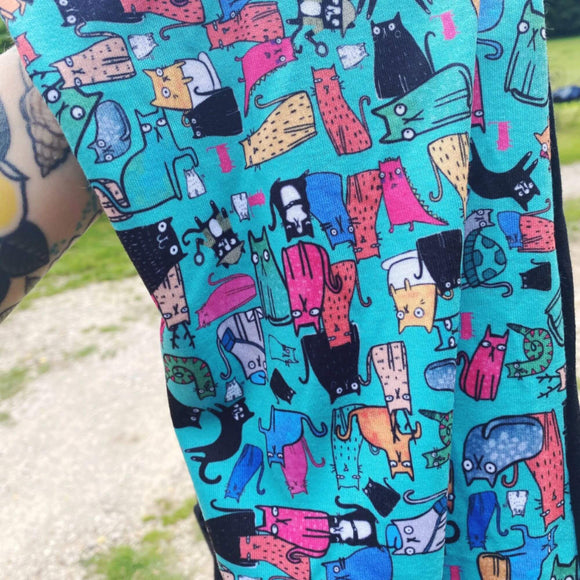 Close-up of a vibrant Katie Abey fabric featuring quirky, colourful cartoon cats in various poses and expressions against a turquoise background. Cats are depicted in bright colours like pink, yellow, orange, and black, with some holding objects or sporting patterns. 