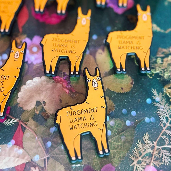 The Judgement Llama Magnet laying on a foliage desk amongst multiple copies of the same magnet. The yellow llama shaped magnet has a scowled expression with black text across its body reading 'judgement llama is watching'.