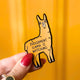 The Judgement Llama Magnet being held up over a yellow background. The yellow llama shaped magnet has a scowled expression with black text across its body reading 'judgement llama is watching'.