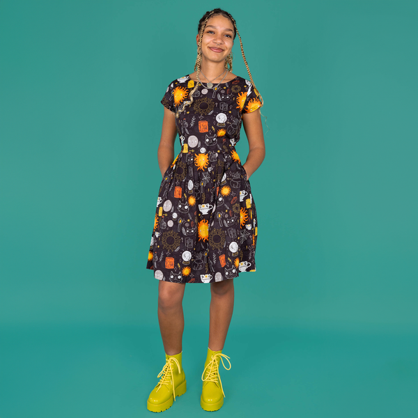 Milly, a femme model with locs, is wearing a black belted tea dress with an all over print of Katie Abey witchy illustrations with a matching fabric belt tied across the waist. The dress has been paired with bright green boots. Milly is smiling at the camera and the background of the photo is a forrest green.