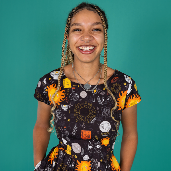 Milly, a femme model with locs, is wearing a black belted tea dress with an all over print of Katie Abey witchy illustrations with a matching fabric belt tied across the waist. The dress has been paired with bright green boots. Milly is smiling at the camera and the background of the photo is a forrest green.