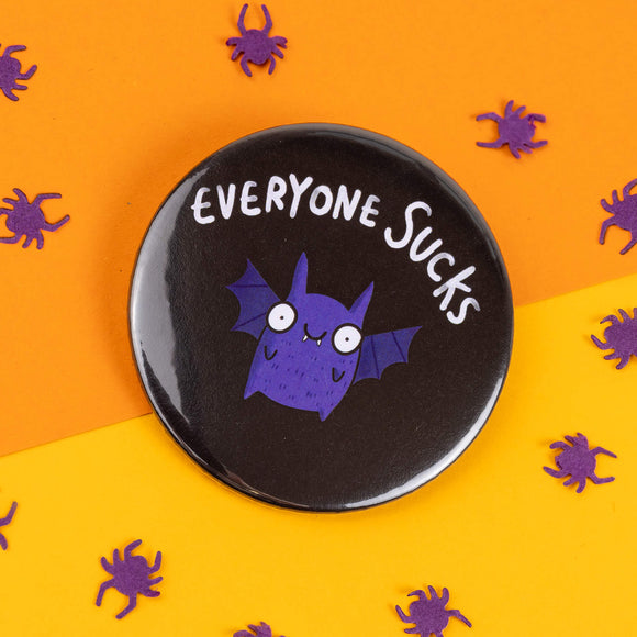 A round black badge featuring a quirky purple bat character with wide eyes and the phrase "Everyone Sucks" in white text at the top. The background is split diagonally between bright orange and yellow, with scattered purple paper spiders. The badge is from Katie Abey, an illustrator who encourages embracing weirdness.