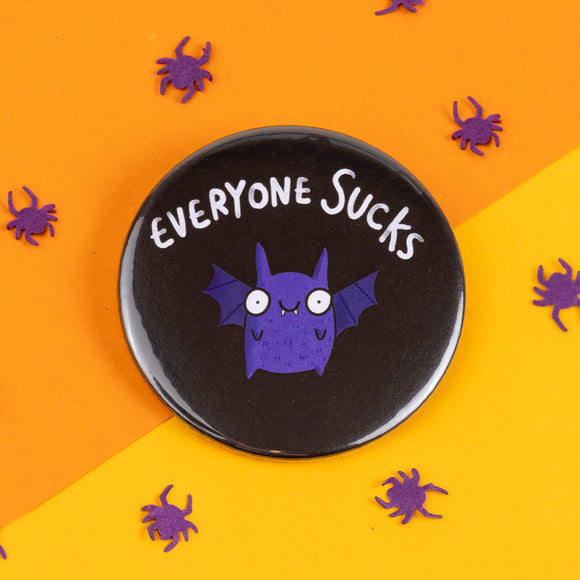 A front view of the black badge featuring a purple bat with "Everyone Sucks" text in white. Purple paper spiders are scattered on a bright orange and yellow background, in line with Katie Abey's quirky, fun style.