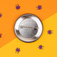 The back of the round metal badge showing its pin clasp. The background is bright orange and yellow with purple paper spiders scattered. This product is from Katie Abey, known for her humour and promoting weirdness.