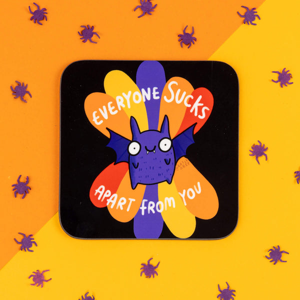 Katie Abey coaster featuring a cute purple bat with wide eyes and tiny fangs, against a vibrant orange and purple petal backdrop. The coaster reads 'Everyone Sucks Apart From You' in white text. The background has orange and yellow tones with small, scattered purple spider confetti. Katie Abey is a small business promoting quirky, weird illustrations that make you smile.