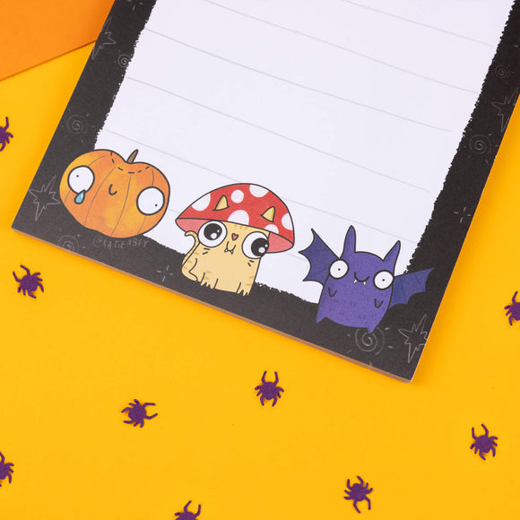 Close-up of a Katie Abey 'Magical and Spooky Musings' notepad. Features a cute cartoon pumpkin, mushroom, and bat character at the bottom of a lined page. Perfect for writing quirky thoughts, with a playful Halloween theme. Designed by an independent illustrator encouraging you to embrace your weirdness.